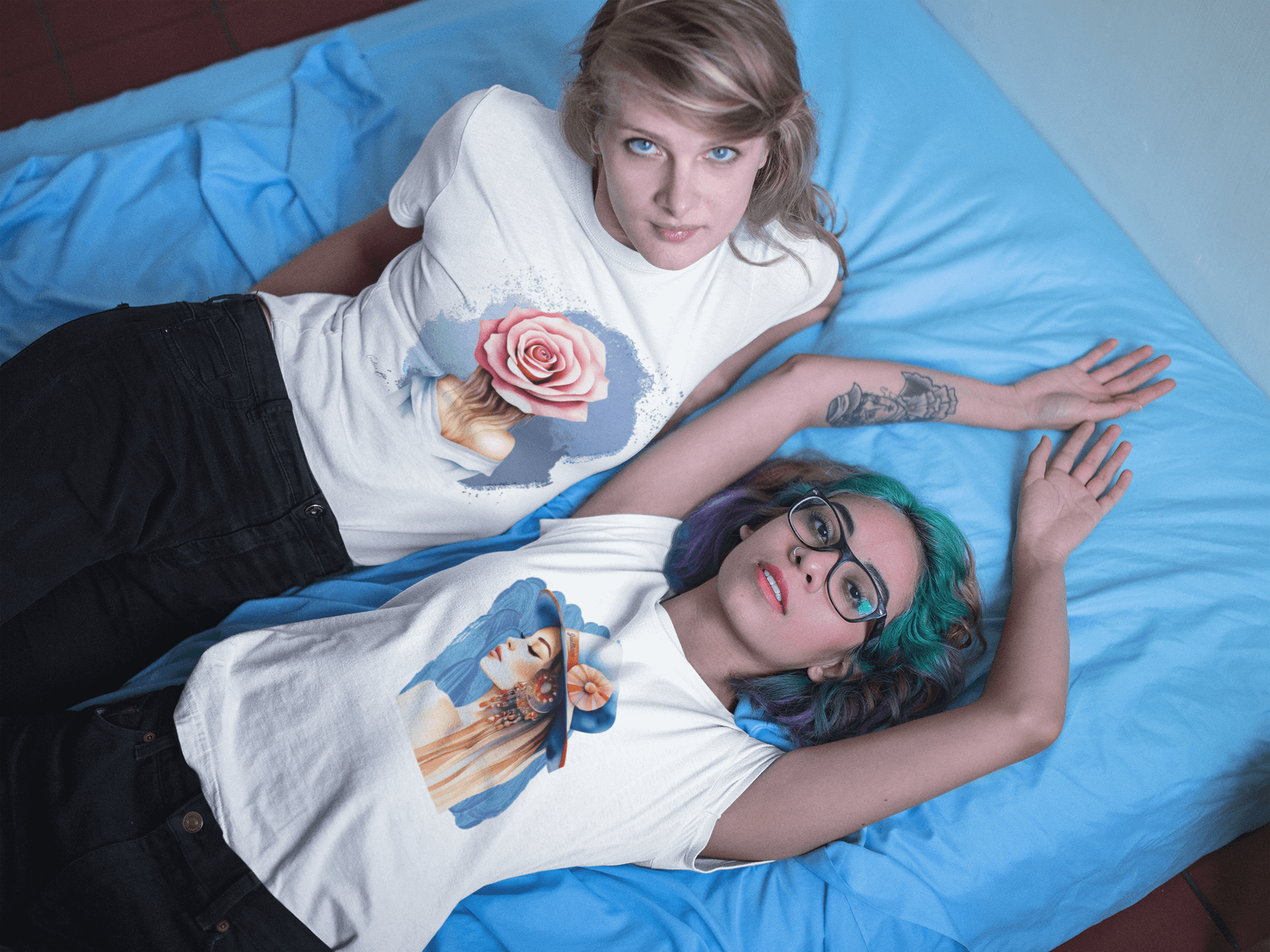 Two women in oversized artistic design t-shirts featuring floral and portrait graphics, lying on a blue bedspread.