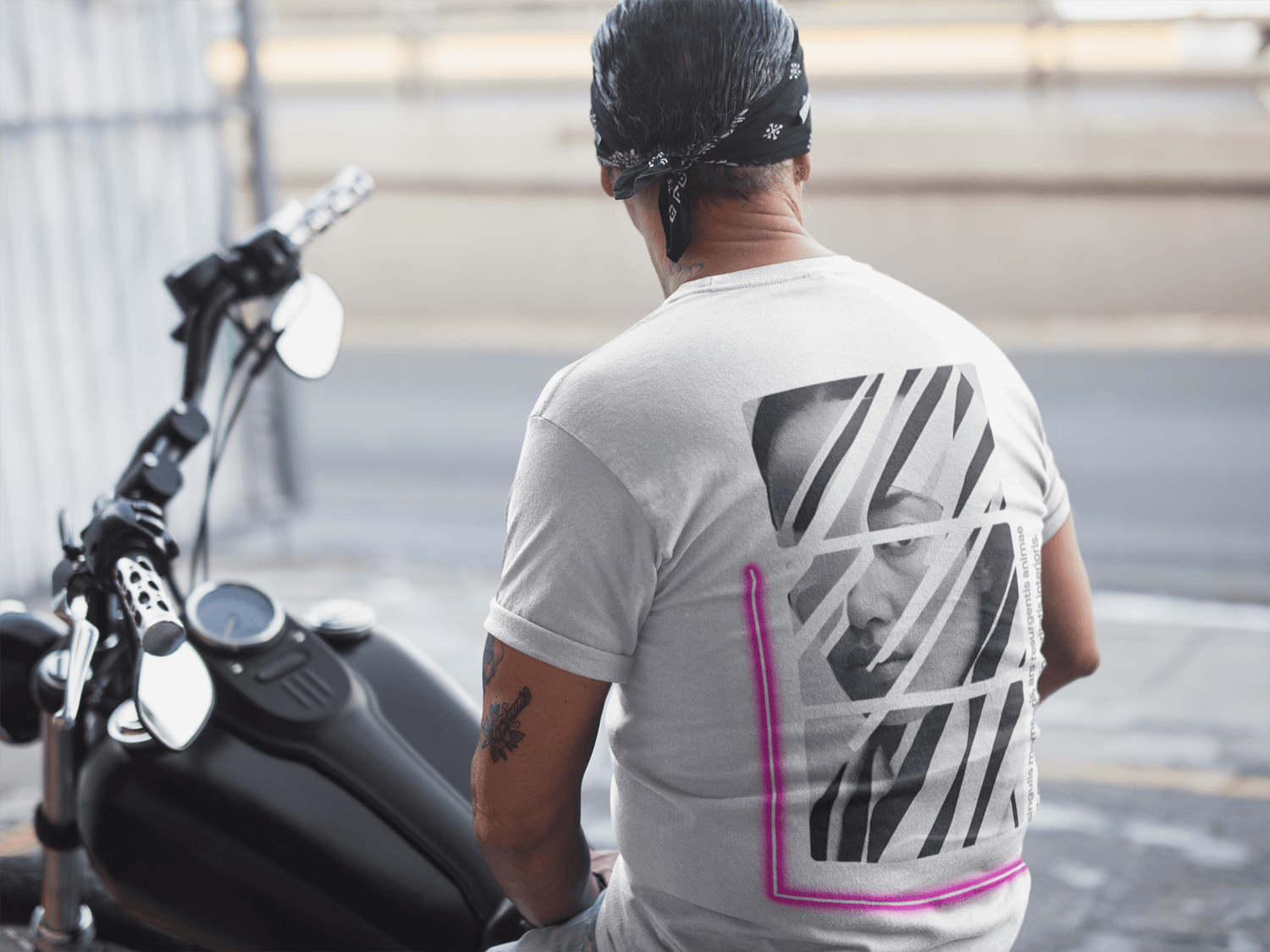 Man wearing a gray oversized neon tee with graphic design, sitting on a motorcycle in an urban setting.